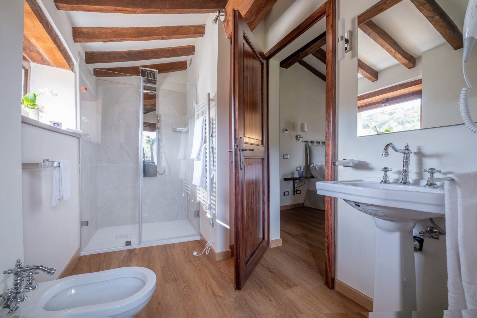 Modern bathroom interior featuring a walk-in shower with glass doors, elegant wooden beams, and stylish fixtures. The space includes a white sink with chrome faucets, a bidet, and a towel rack, complemented by warm wooden flooring and natural light from a window. | Only on SopranoVillas