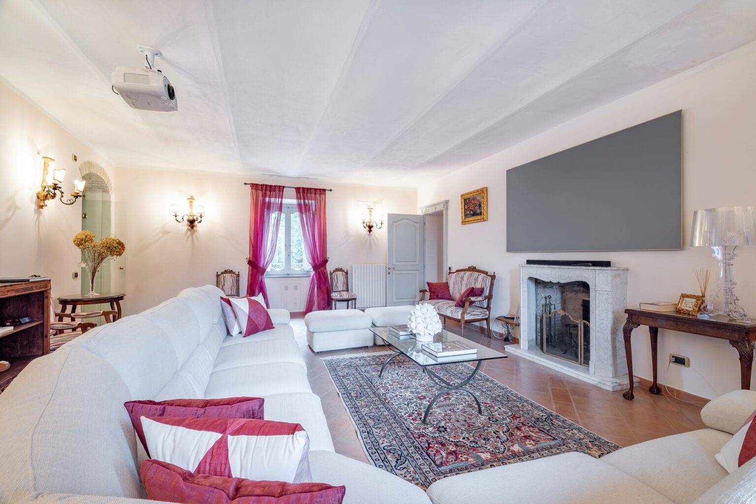 Spacious and elegantly designed living room featuring a comfortable white sectional sofa with decorative pillows, a stylish glass coffee table, and a vintage fireplace. Natural light streams through large windows adorned with sheer red curtains, complemented by decorative wall sconces. A projector is mounted on the ceiling, and a large flat-screen TV is positioned above the fireplace, creating a cozy entertainment space. | Only on SopranoVillas