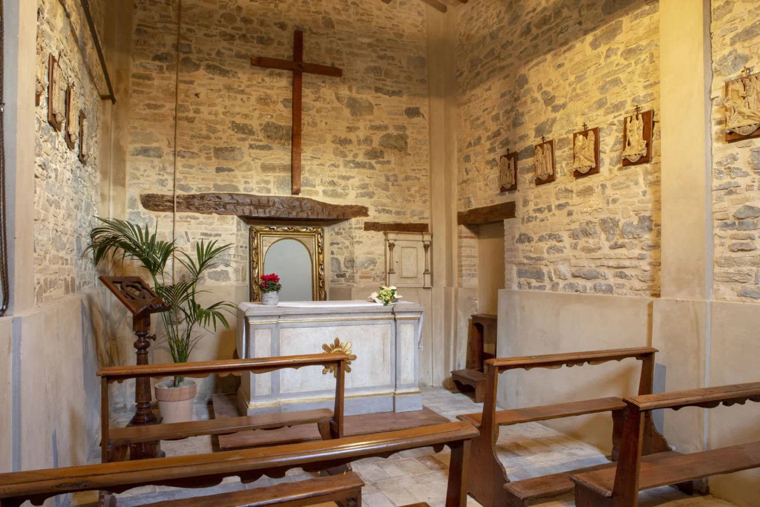 Interior of a small chapel featuring a wooden cross, stone walls, and a simple altar adorned with flowers. The space includes wooden benches for seating and a decorative mirror above the altar, creating a serene atmosphere for worship. | Only on SopranoVillas