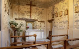 Interior of a small chapel featuring a wooden cross, stone walls, and a simple altar adorned with flowers. The space includes wooden benches for seating and a decorative mirror above the altar, creating a serene atmosphere for worship.