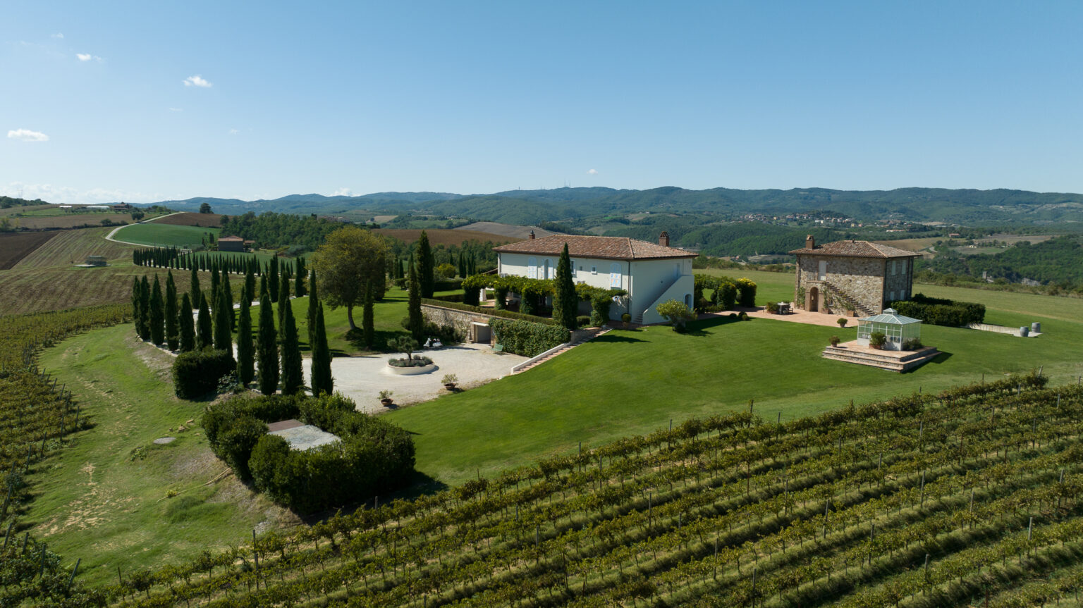 Aerial view of a picturesque Tuscan villa surrounded by lush green vineyards and cypress trees, with rolling hills in the background under a clear blue sky. | Only on SopranoVillas