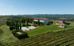 Aerial view of a picturesque Tuscan villa surrounded by lush green vineyards and cypress trees, with rolling hills in the background under a clear blue sky.