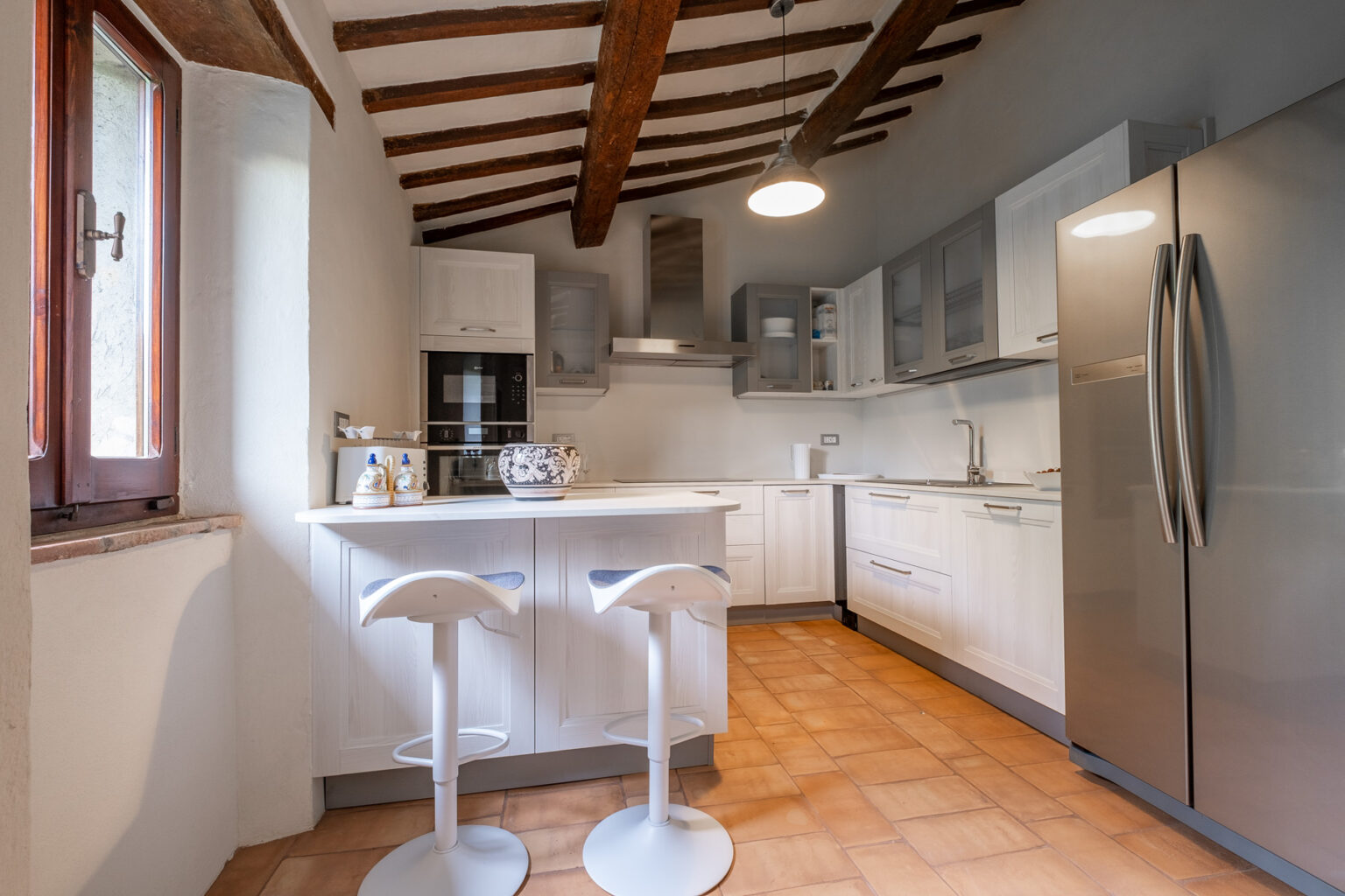 Modern kitchen interior featuring white cabinetry, stainless steel appliances, and a cozy breakfast bar with two stools. Exposed wooden beams and a large window add character and natural light to the space. The warm tile flooring complements the contemporary design. | Only on SopranoVillas