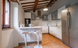 Modern kitchen interior featuring white cabinetry, stainless steel appliances, and a cozy breakfast bar with two stools. Exposed wooden beams and a large window add character and natural light to the space. The warm tile flooring complements the contemporary design.
