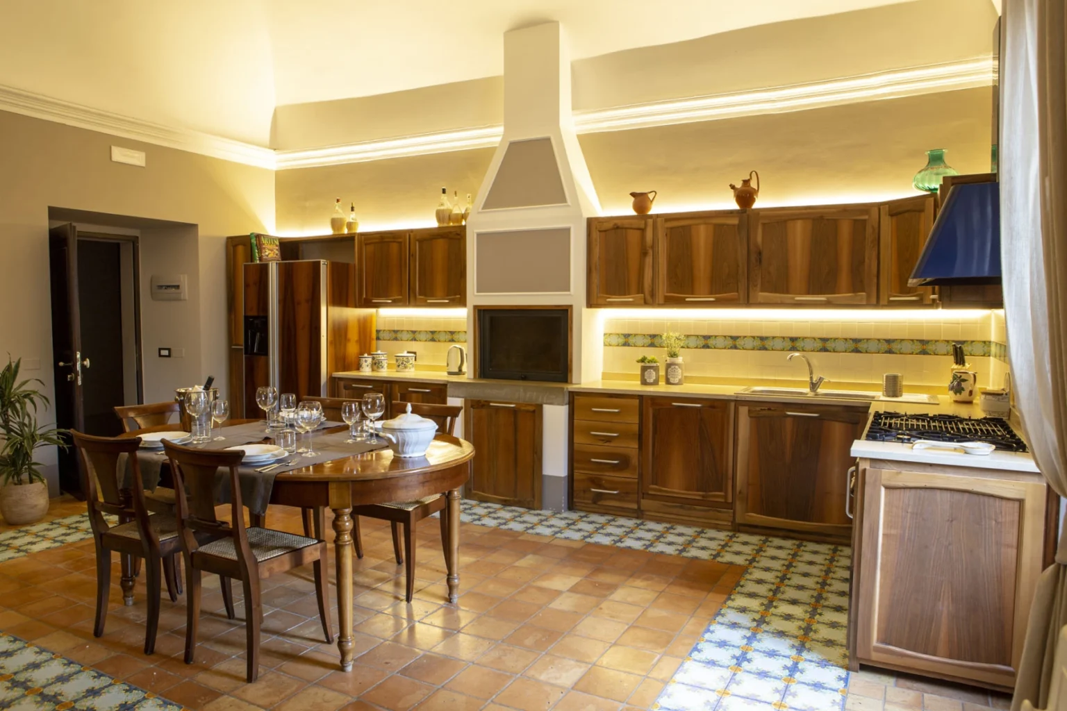 Spacious modern kitchen featuring wooden cabinetry, a dining table set for four, and a colorful tiled floor. The kitchen is well-lit with ambient lighting and includes stainless steel appliances, a gas stove, and decorative elements on the shelves. | Only on SopranoVillas