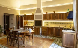 Spacious modern kitchen featuring wooden cabinetry, a dining table set for four, and a colorful tiled floor. The kitchen is well-lit with ambient lighting and includes stainless steel appliances, a gas stove, and decorative elements on the shelves.