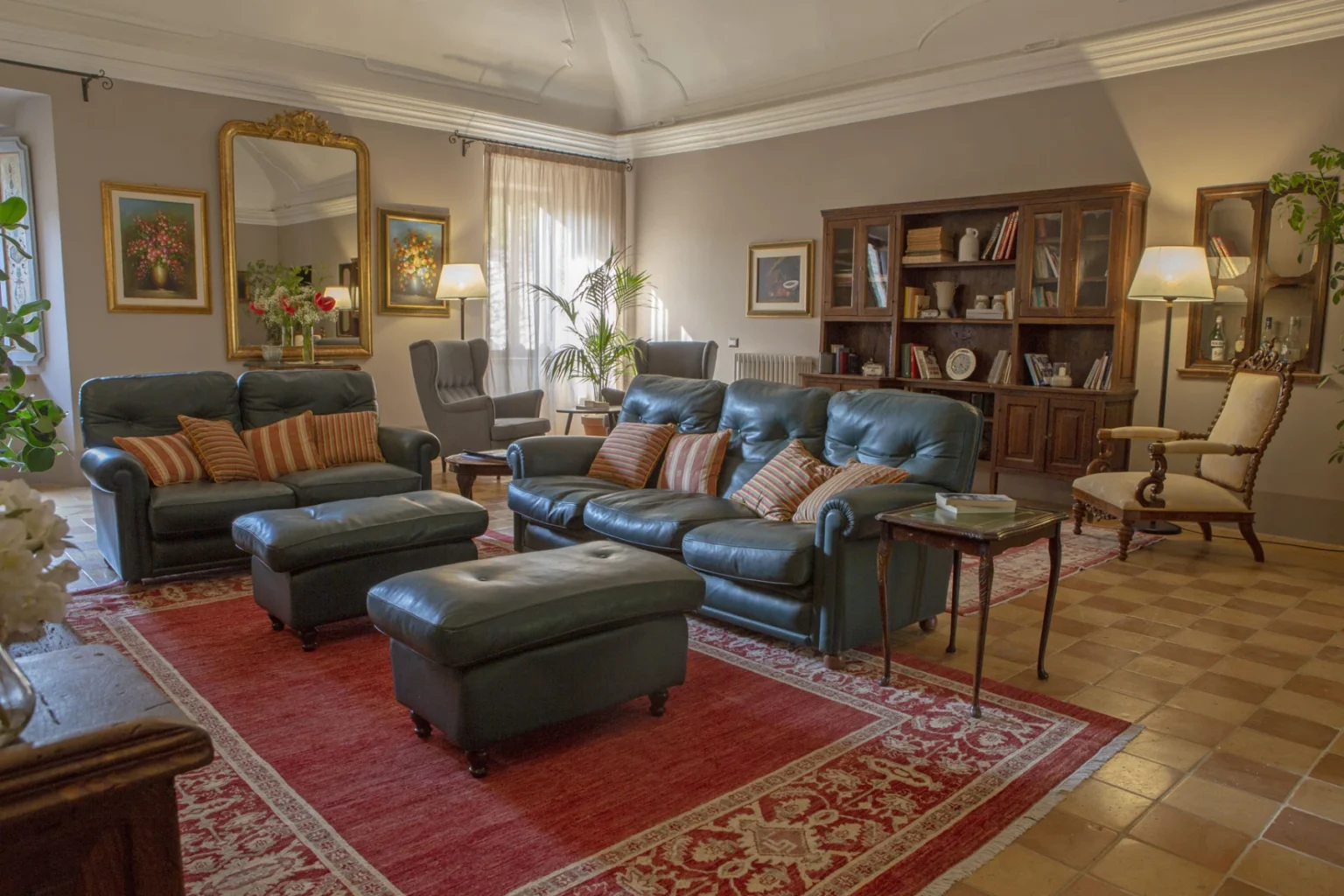 Cozy living room featuring dark leather sofas with striped cushions, an ornate mirror, decorative paintings, a wooden bookshelf filled with books, and a warm lighting ambiance. The space is enhanced by a red area rug and indoor plants, creating a welcoming atmosphere. | Only on SopranoVillas