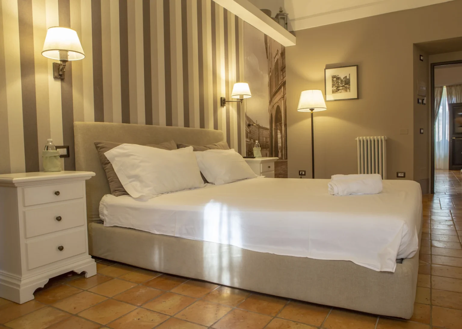 Cozy bedroom featuring a comfortable bed with white linens, decorative pillows, and stylish bedside lamps. The room has a warm color palette with striped wallpaper, a small nightstand, and elegant flooring. Ideal for relaxation and restful stays. | Only on SopranoVillas