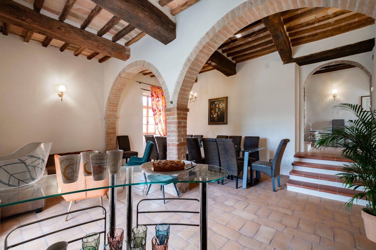 Spacious and elegantly designed dining area featuring arched brick accents and wooden beams, with a glass table displaying colorful glassware and a dessert, surrounded by stylish seating and a vibrant window treatment. Ideal for showcasing a cozy yet sophisticated interior space. | Only on SopranoVillas