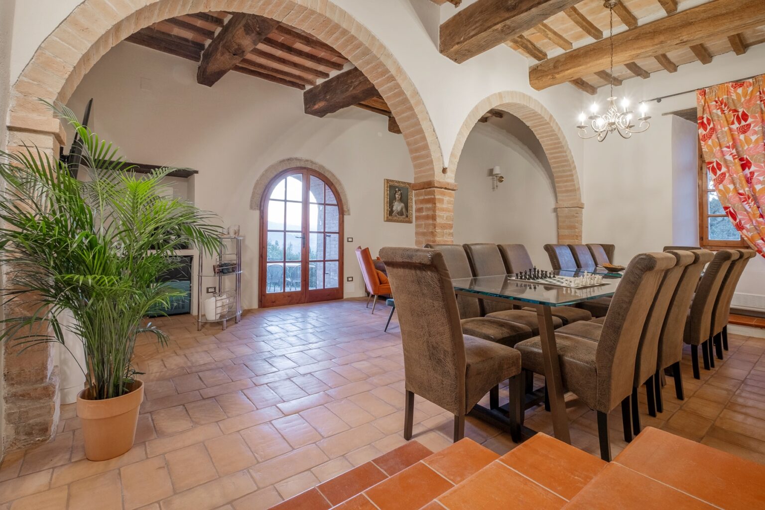 Spacious dining area featuring a large glass table surrounded by upholstered chairs, rustic wooden beams, and arched doorways. A potted plant adds greenery, while large windows provide natural light and views of the outdoors. | Only on SopranoVillas