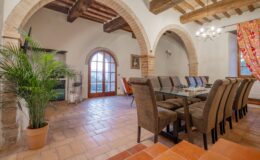 Spacious dining area featuring a large glass table surrounded by upholstered chairs, rustic wooden beams, and arched doorways. A potted plant adds greenery, while large windows provide natural light and views of the outdoors.
