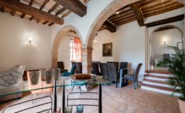 Spacious and elegantly designed dining area featuring arched brick accents and wooden beams, with a glass table displaying colorful glassware and a dessert, surrounded by stylish seating and a vibrant window treatment. Ideal for showcasing a cozy yet sophisticated interior space.