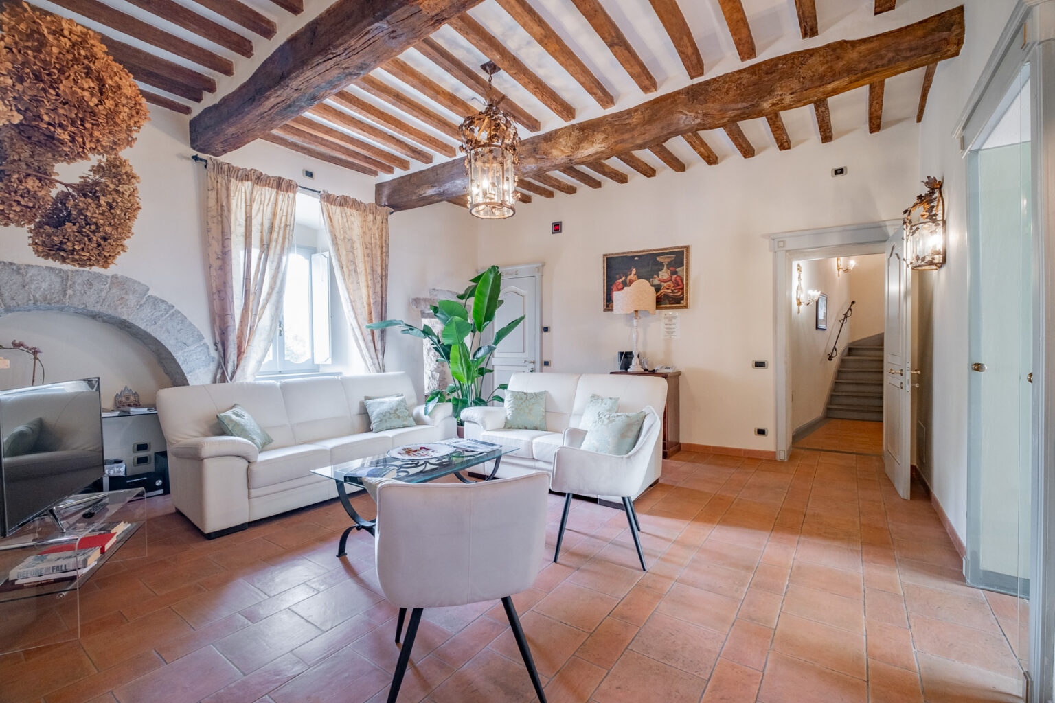 Spacious living room featuring rustic wooden beams, elegant furnishings, and large windows. The area includes a white leather sofa, glass coffee table, and decorative plants, creating a cozy and inviting atmosphere. | Only on SopranoVillas