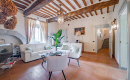 Spacious living room featuring rustic wooden beams, elegant furnishings, and large windows. The area includes a white leather sofa, glass coffee table, and decorative plants, creating a cozy and inviting atmosphere.