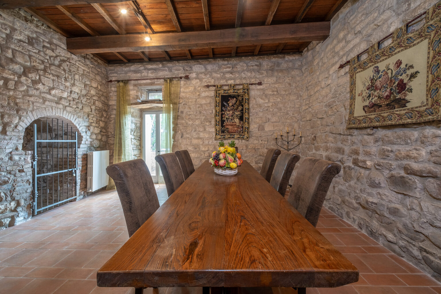 Rustic dining room featuring a large wooden table surrounded by upholstered chairs, with stone walls and decorative tapestries. Natural light streams through glass doors, enhancing the warm ambiance of the space. A centerpiece of fresh fruit adds a touch of color to the table. | Only on SopranoVillas