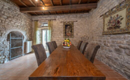Rustic dining room featuring a large wooden table surrounded by upholstered chairs, with stone walls and decorative tapestries. Natural light streams through glass doors, enhancing the warm ambiance of the space. A centerpiece of fresh fruit adds a touch of color to the table.