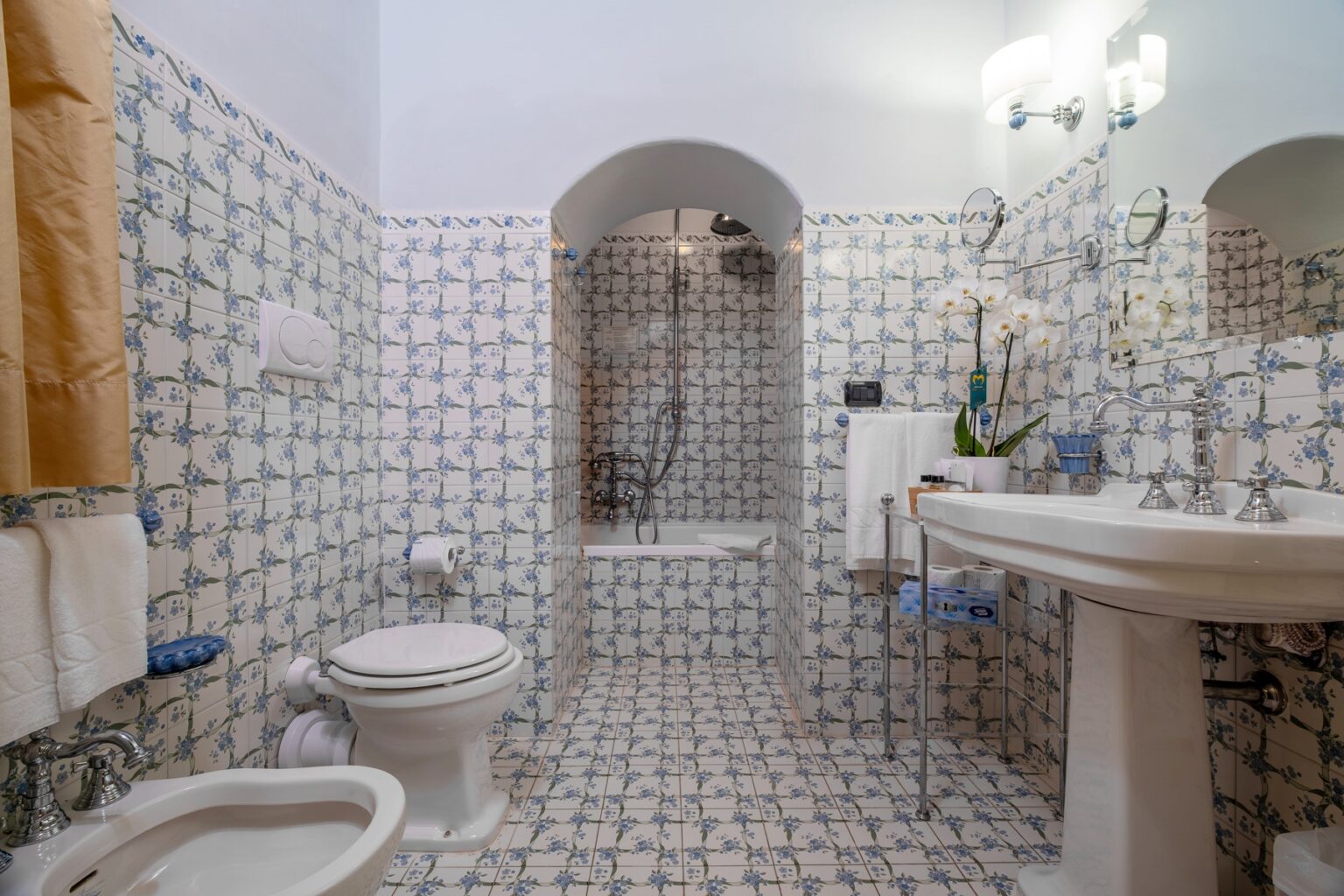 Modern bathroom featuring floral patterned tiles, a bathtub with a shower, a white pedestal sink, and elegant fixtures. The space is well-lit with a mirror and decorative plants, creating a fresh and inviting atmosphere. | Only on SopranoVillas