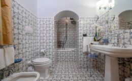 Modern bathroom featuring floral patterned tiles, a bathtub with a shower, a white pedestal sink, and elegant fixtures. The space is well-lit with a mirror and decorative plants, creating a fresh and inviting atmosphere.