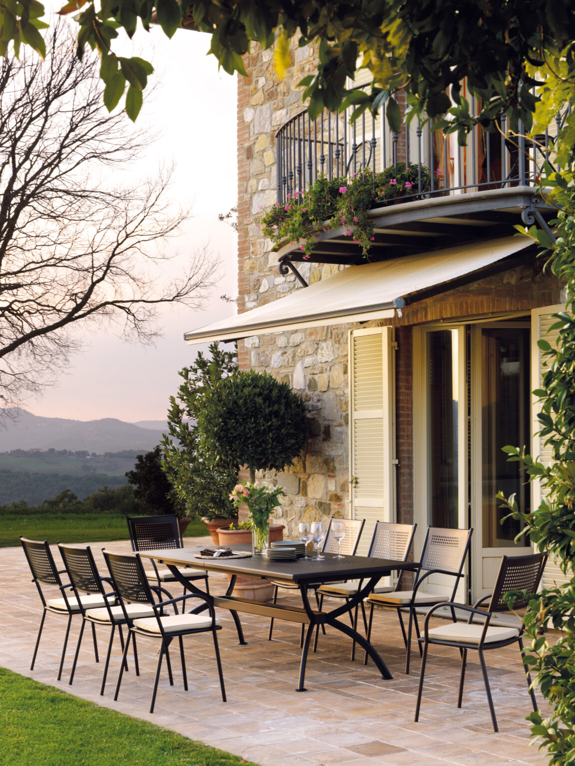 Outdoor dining area featuring a stylish table and chairs set against a stone wall, with a balcony adorned with flowers. The scene captures a serene landscape, perfect for al fresco dining in a tranquil setting. | Only on SopranoVillas