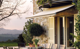 Outdoor dining area featuring a stylish table and chairs set against a stone wall, with a balcony adorned with flowers. The scene captures a serene landscape, perfect for al fresco dining in a tranquil setting.
