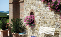 Charming outdoor patio featuring a stone wall adorned with vibrant pink flowers, a white metal bench, and two small tables. Ideal for relaxation, the setting includes a view of lush greenery and a clear blue sky, perfect for enjoying sunny days.