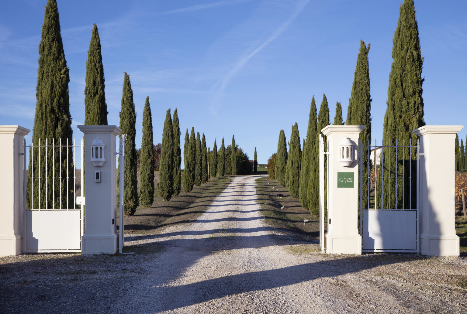 Gravel driveway leading to an elegant entrance gate flanked by tall cypress trees, under a clear blue sky. The gate features a sign reading "La Ville," inviting visitors to a picturesque estate. | Only on SopranoVillas