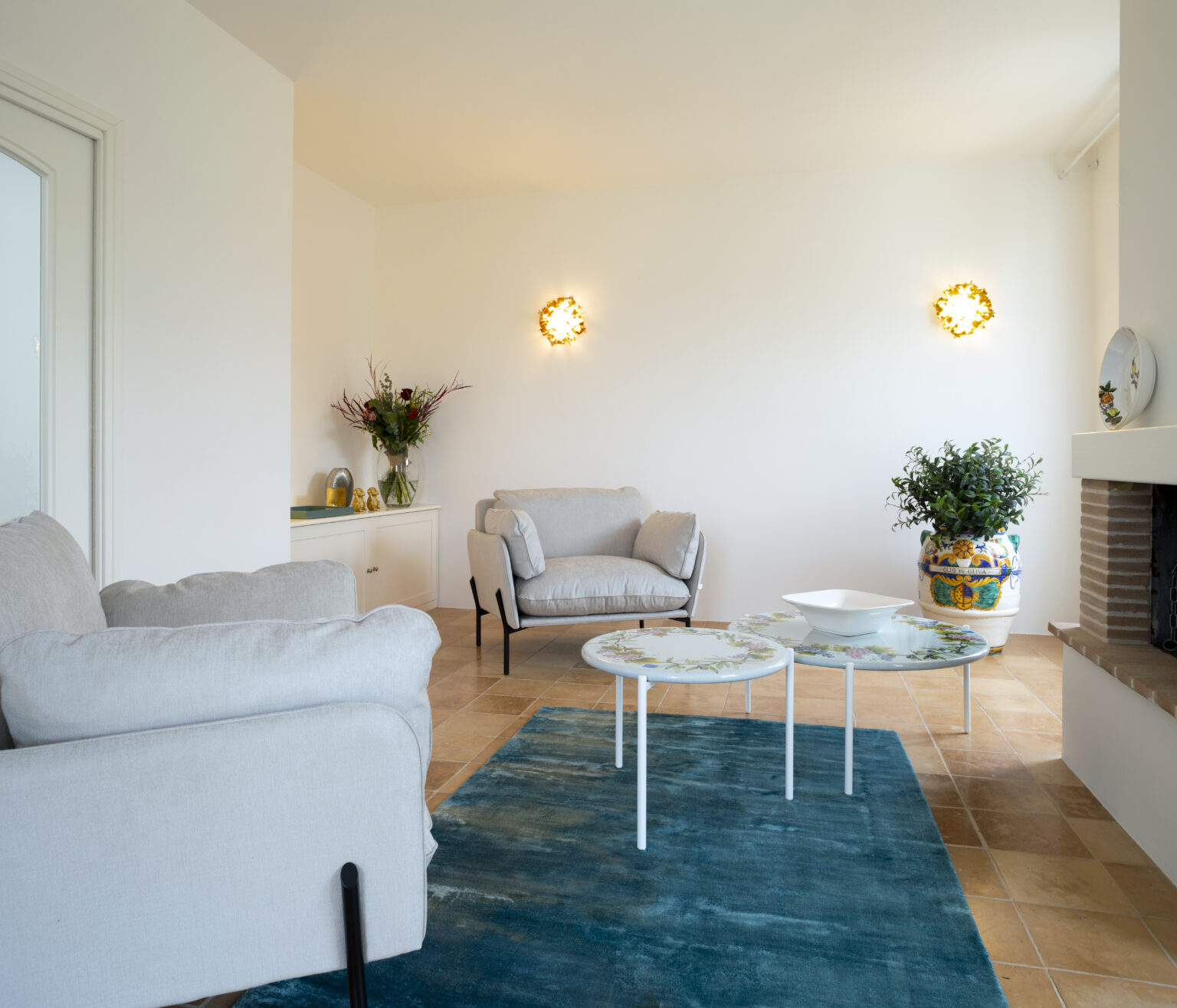 Bright and modern living room featuring two light gray armchairs, a decorative coffee table with floral design, and a vibrant blue area rug. Natural light floods the space through a nearby window, highlighting a stylish wall lamp and a potted plant. A cozy ambiance is created by a neutral color palette and elegant decor. | Only on SopranoVillas