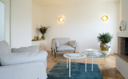 Bright and modern living room featuring two light gray armchairs, a decorative coffee table with floral design, and a vibrant blue area rug. Natural light floods the space through a nearby window, highlighting a stylish wall lamp and a potted plant. A cozy ambiance is created by a neutral color palette and elegant decor.