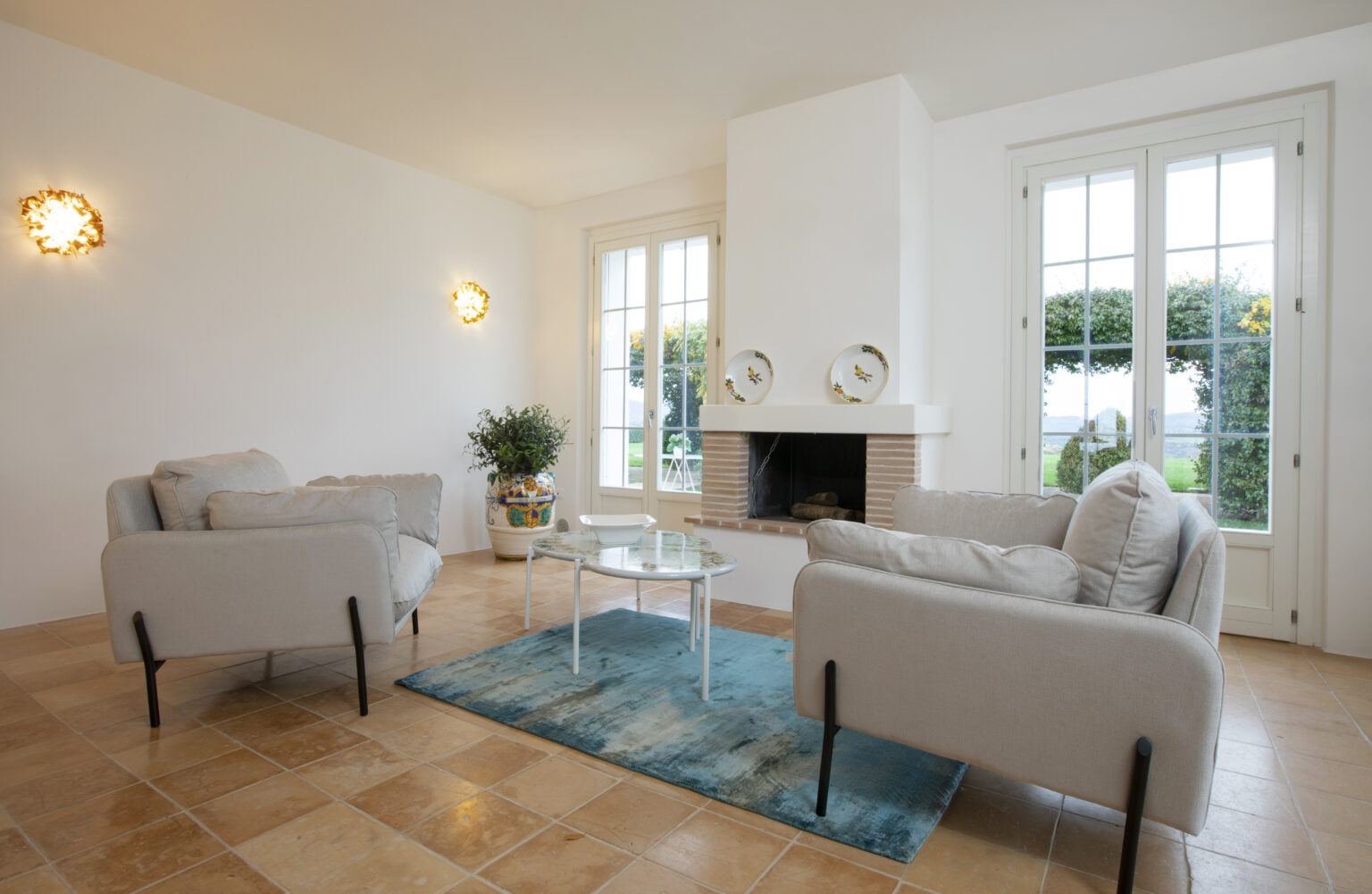 Cozy living room featuring two light gray sofas, a glass coffee table, and a decorative rug. The space includes a modern fireplace and large windows that provide natural light and a view of the garden. Elegant wall sconces add a warm ambiance to the room. | Only on SopranoVillas