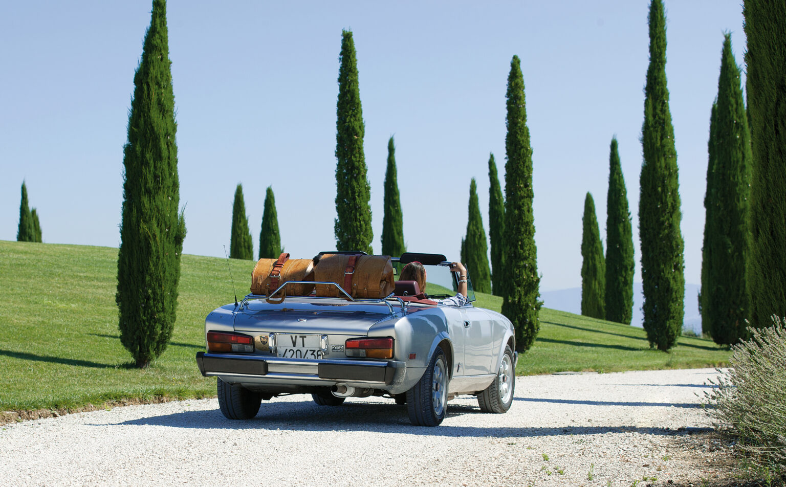 Classic convertible car parked on a gravel road surrounded by tall cypress trees and lush green grass, suggesting a scenic countryside getaway. The car features vintage luggage on the back, enhancing the travel theme. | Only on SopranoVillas