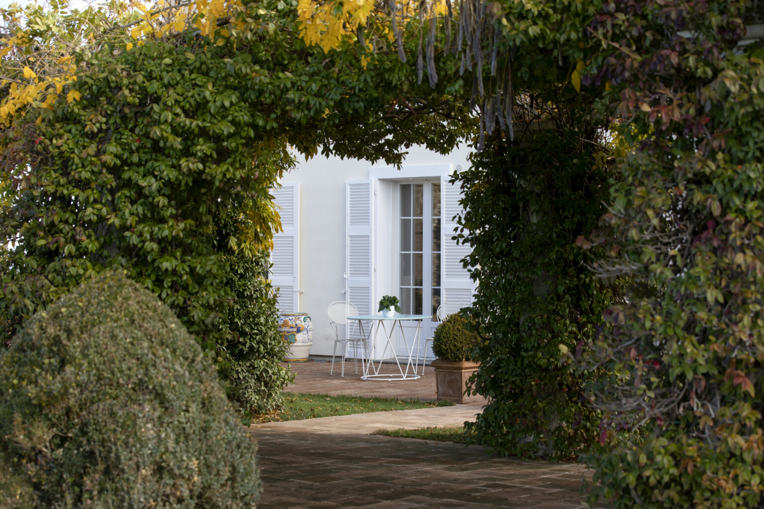 Charming outdoor space framed by lush greenery, featuring a small white table and chairs set on a stone patio, with a decorative pot and a view of a house with white shutters in the background. | Only on SopranoVillas
