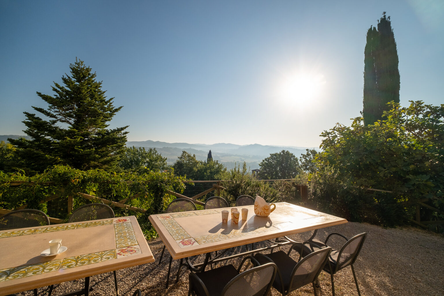 Scenic outdoor dining area with a table set for coffee, overlooking a picturesque landscape of hills and trees under a bright sun. | Only on SopranoVillas
