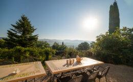 Scenic outdoor dining area with a table set for coffee, overlooking a picturesque landscape of hills and trees under a bright sun.