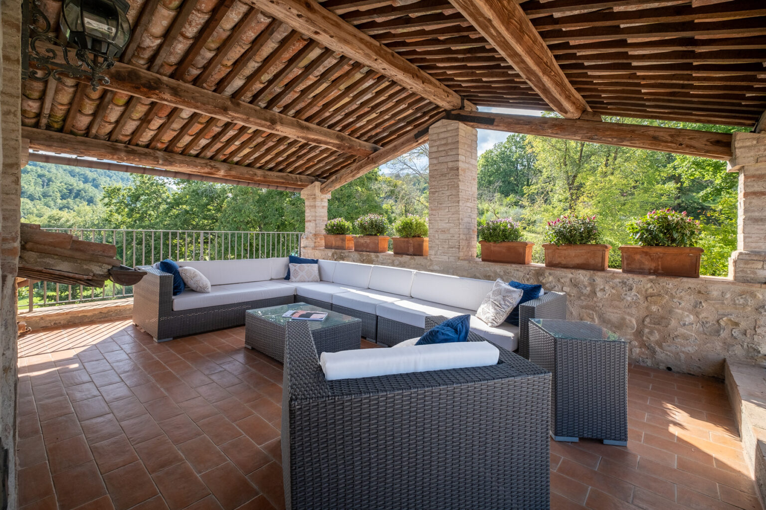Spacious outdoor terrace featuring a comfortable seating area with a sectional sofa, coffee table, and decorative cushions. The area is shaded by a wooden roof and surrounded by lush greenery and potted plants, creating a serene and inviting atmosphere. | Only on SopranoVillas