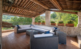 Spacious outdoor terrace featuring a comfortable seating area with a sectional sofa, coffee table, and decorative cushions. The area is shaded by a wooden roof and surrounded by lush greenery and potted plants, creating a serene and inviting atmosphere.