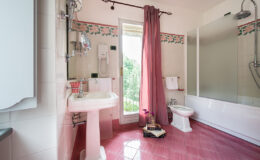 Bright and airy bathroom featuring a pink tiled floor, a white pedestal sink, and a shower area with a glass partition. Decorative floral accents adorn the walls, complemented by a long pink curtain and a small decorative table with flowers. Natural light streams in from a window, enhancing the inviting atmosphere.