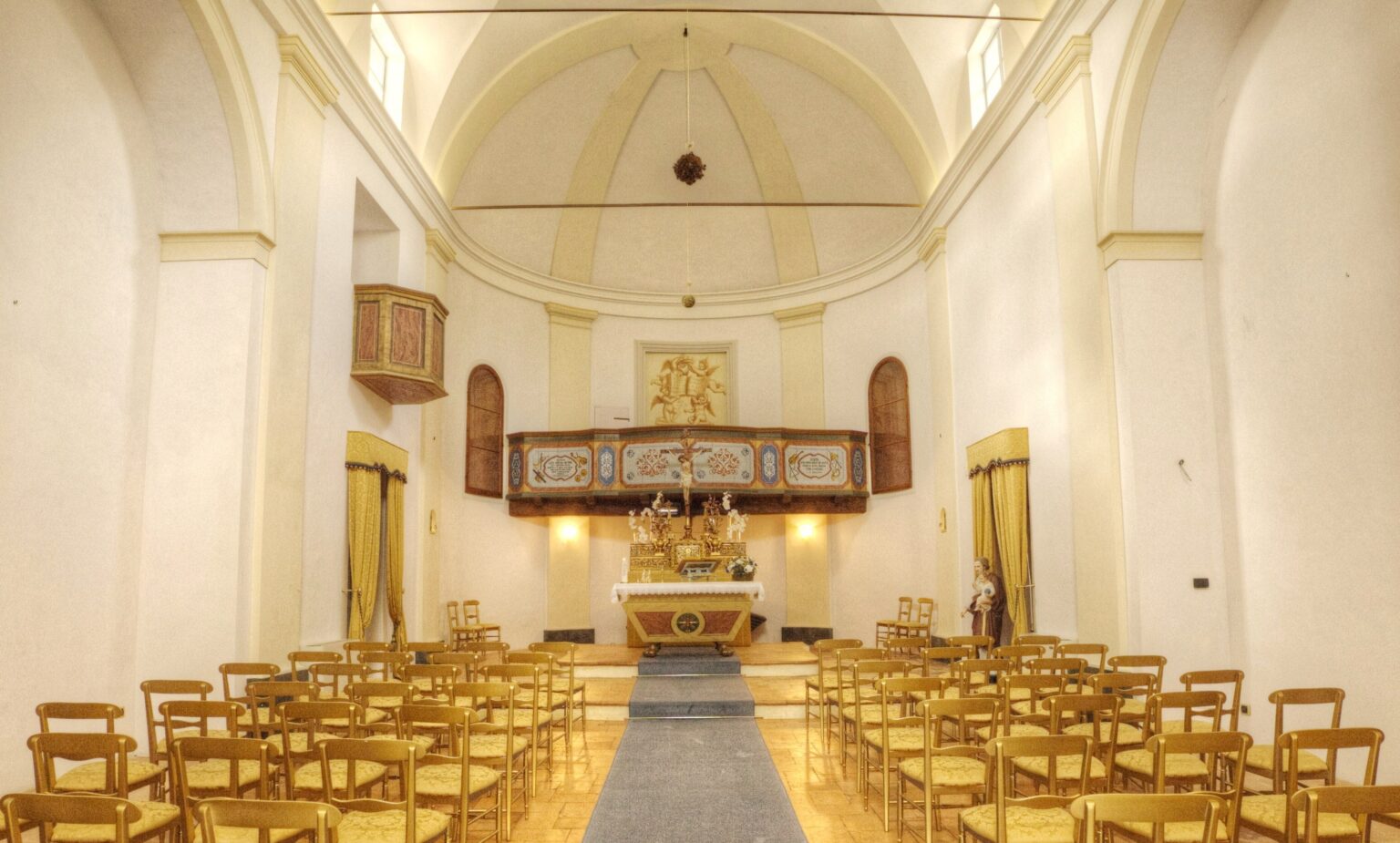Interior view of a beautifully designed church, featuring rows of golden chairs, an altar adorned with floral arrangements, and a wooden balcony with decorative elements. The soft lighting and elegant architecture create a serene atmosphere, suitable for worship and gatherings. | Only on SopranoVillas