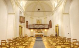 Interior view of a beautifully designed church, featuring rows of golden chairs, an altar adorned with floral arrangements, and a wooden balcony with decorative elements. The soft lighting and elegant architecture create a serene atmosphere, suitable for worship and gatherings.