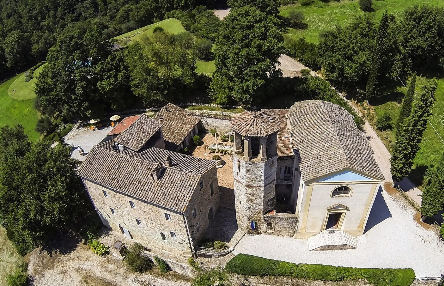 Aerial view of a historic stone villa complex with a bell tower, surrounded by lush greenery and picturesque landscapes, showcasing traditional architecture and a charming courtyard. | Only on SopranoVillas