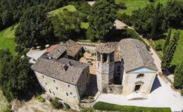 Aerial view of a historic stone villa complex with a bell tower, surrounded by lush greenery and picturesque landscapes, showcasing traditional architecture and a charming courtyard.