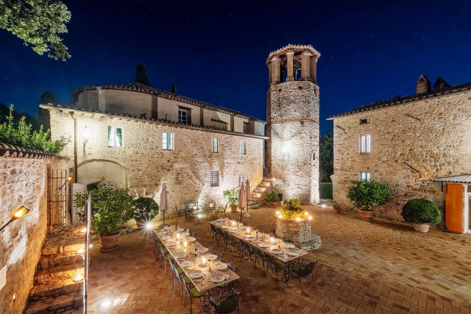 Illuminated outdoor dining area featuring a long table set for a meal, surrounded by stone buildings and a historic tower at night, showcasing a picturesque Italian villa landscape. | Only on SopranoVillas
