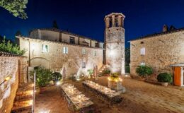 Illuminated outdoor dining area featuring a long table set for a meal, surrounded by stone buildings and a historic tower at night, showcasing a picturesque Italian villa landscape.