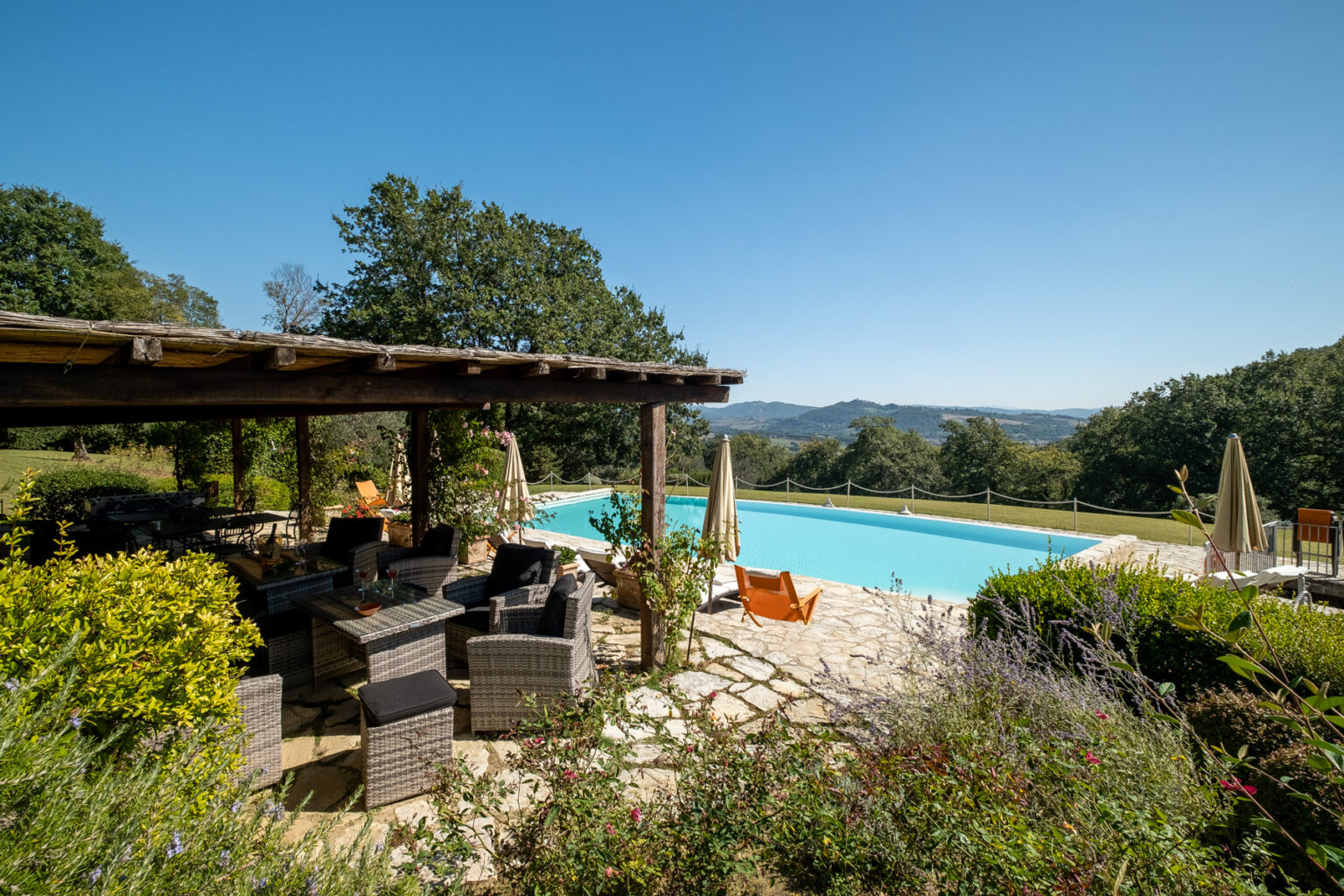 Scenic view of a luxury outdoor pool area surrounded by lush greenery, featuring a shaded seating area with wicker furniture and colorful flowers in the foreground. The clear blue sky and distant hills create a tranquil atmosphere perfect for relaxation. | Only on SopranoVillas