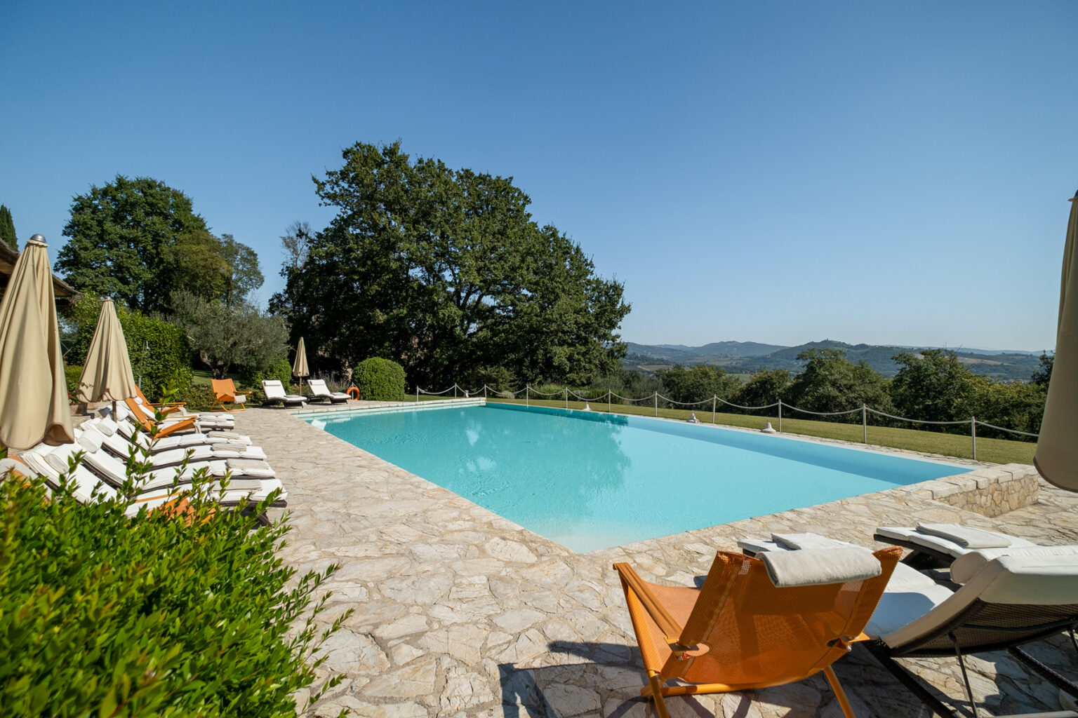 Scenic outdoor swimming pool surrounded by lounge chairs and umbrellas, set against a backdrop of lush greenery and rolling hills under a clear blue sky. Ideal for relaxation and leisure. | Only on SopranoVillas
