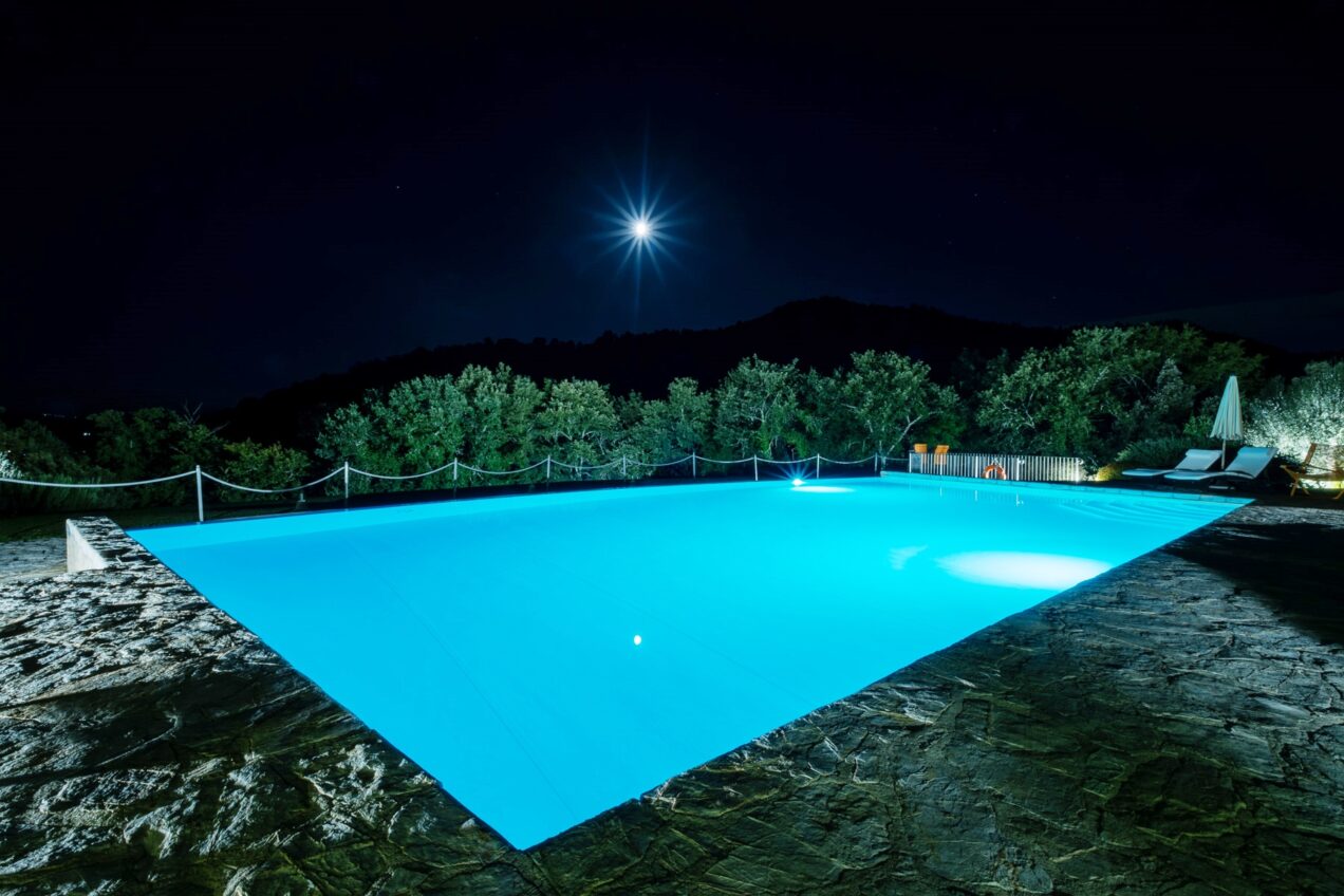 Illuminated swimming pool at night surrounded by lush greenery and a starry sky, featuring a clear blue water surface and stone deck, creating a serene and luxurious atmosphere.