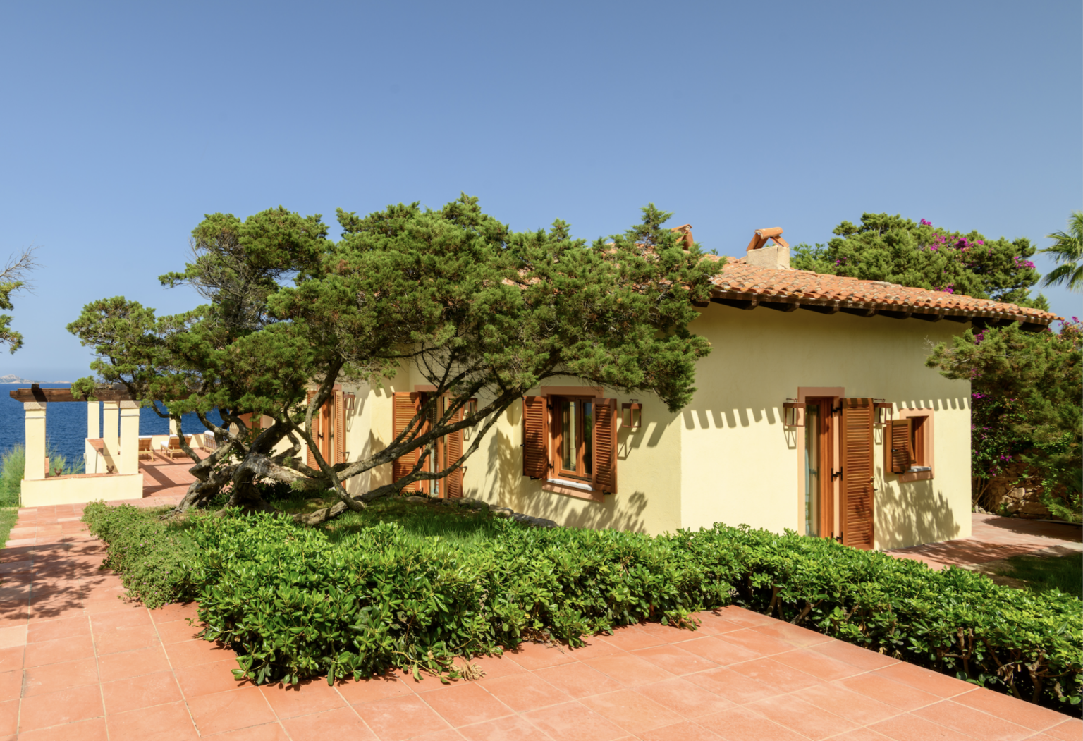 Charming seaside cottage surrounded by lush greenery and vibrant flowers, featuring rustic wooden windows and a tiled roof, with a clear blue ocean visible in the background. | Only on SopranoVillas