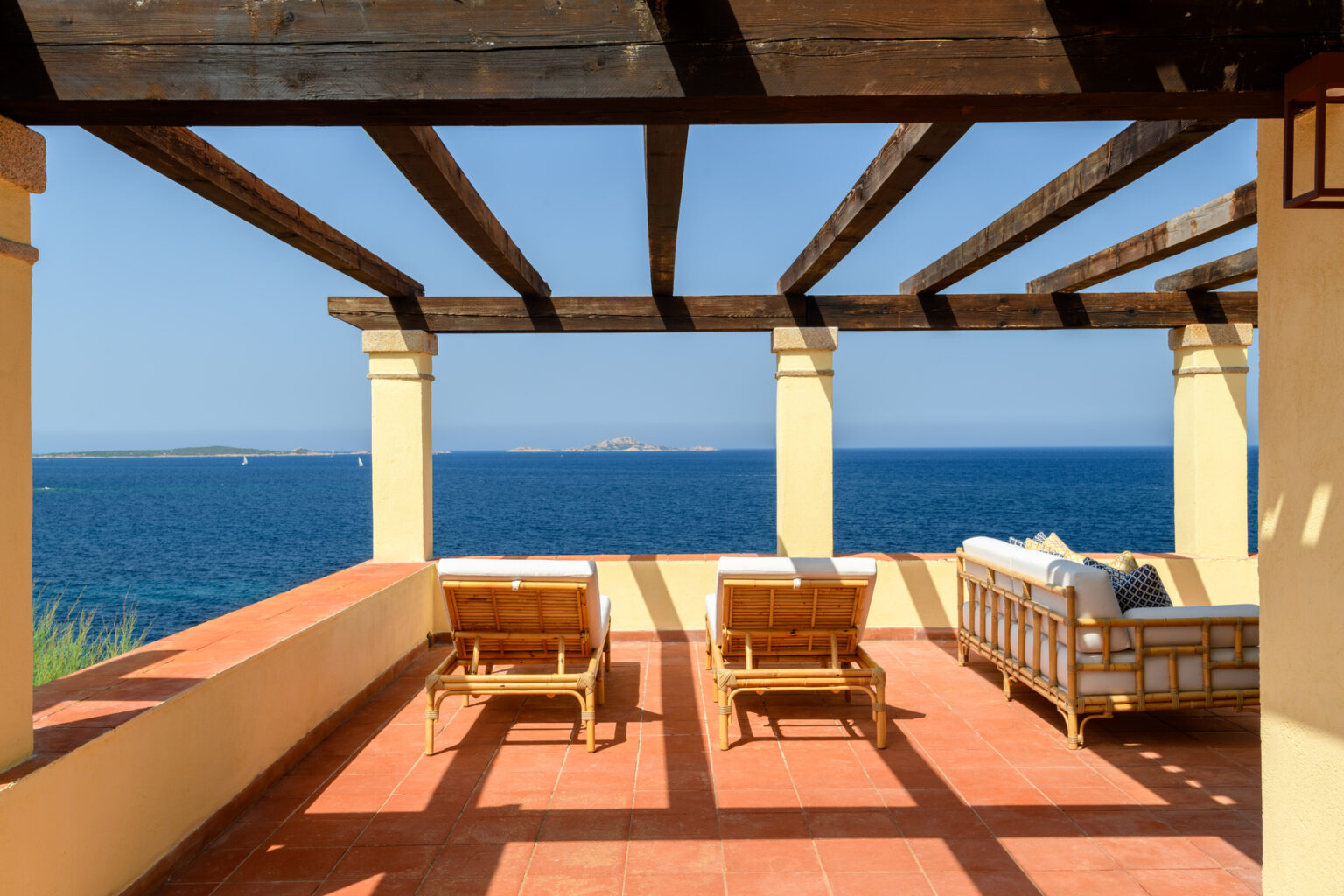 Cozy outdoor terrace with wooden lounge chairs overlooking a serene blue ocean, framed by a rustic pergola and bright sunlight, ideal for relaxation and enjoying coastal views. | Only on SopranoVillas