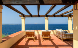 Cozy outdoor terrace with wooden lounge chairs overlooking a serene blue ocean, framed by a rustic pergola and bright sunlight, ideal for relaxation and enjoying coastal views.