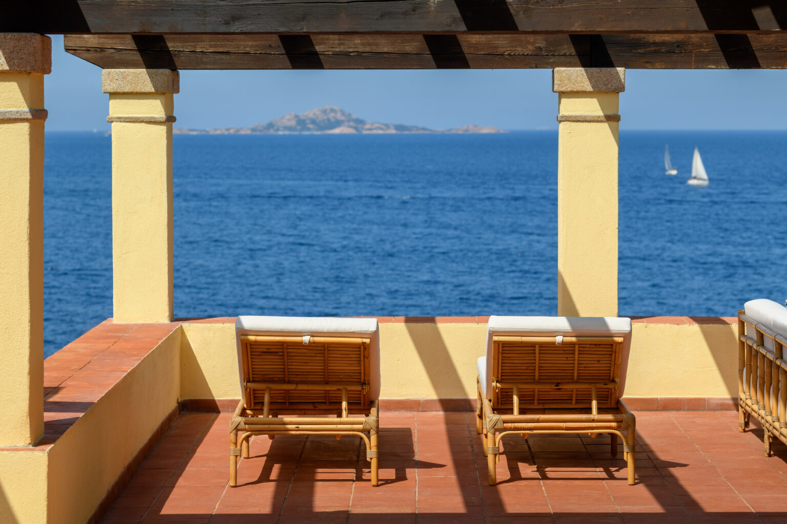 Cozy outdoor terrace with two lounge chairs overlooking a serene ocean view and distant sailboats, framed by elegant columns and a clear blue sky. | Only on SopranoVillas