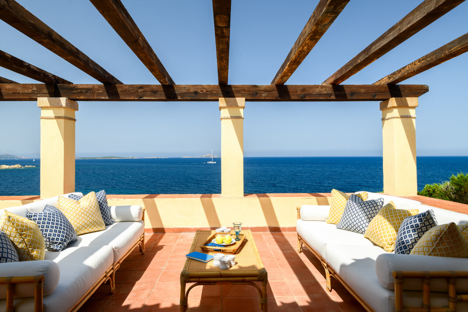 Cozy outdoor seating area with decorative pillows overlooking a serene blue sea, featuring a wooden pergola and a table with refreshments. Ideal for relaxation and enjoying coastal views. | Only on SopranoVillas